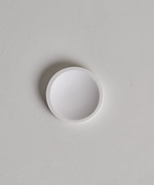 A Coast Soap Dish, small, round, and white, rests on a light gray textured surface with a faint shadow on its left side.