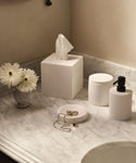 A marble bathroom countertop displays the Coast Soap Dish, handcasted resin and made in Mexico, next to a vase of white flowers, a white tissue box, a soap dispenser, a jar, and a jewelry dish holding a necklace and rings.