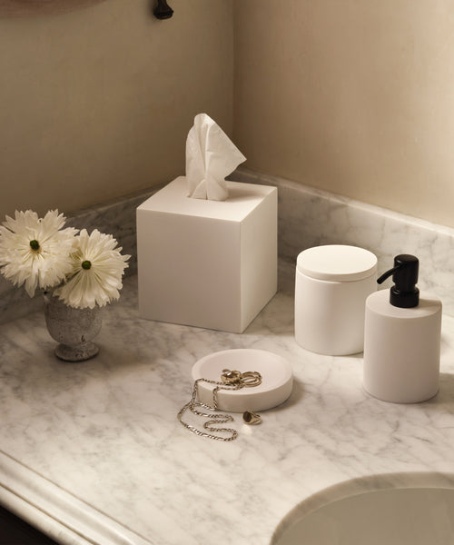 A marble bathroom countertop displays the Coast Soap Dish, handcasted resin and made in Mexico, next to a vase of white flowers, a white tissue box, a soap dispenser, a jar, and a jewelry dish holding a necklace and rings.