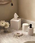 A marble bathroom countertop displays the Coast Soap Dispenser, a white tissue box, container, round tray with jewelry, and a small vase of white daisies. Soft lighting and minimal decor highlight handcast resin accents made in Mexico.