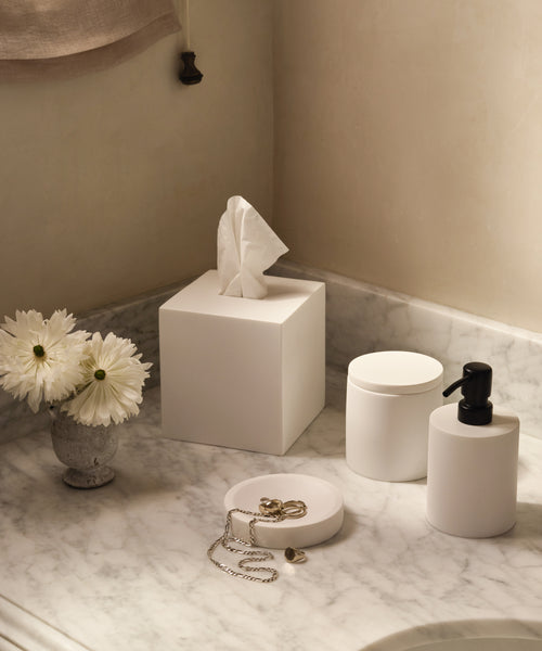 A marble bathroom countertop displays the Coast Soap Dispenser, a white tissue box, container, round tray with jewelry, and a small vase of white daisies. Soft lighting and minimal decor highlight handcast resin accents made in Mexico.