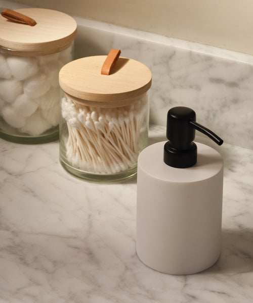 A Coast Soap Dispenser sits on a marble bathroom counter beside two glass jars with wooden lids—one containing cotton balls and the other cotton swabs.