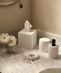 A marble bathroom countertop displays elegant home accessories: white containers, the Coast Tissue Cover, a soap dispenser, a small vase with white flowers, and jewelry including a chain necklace and rings.