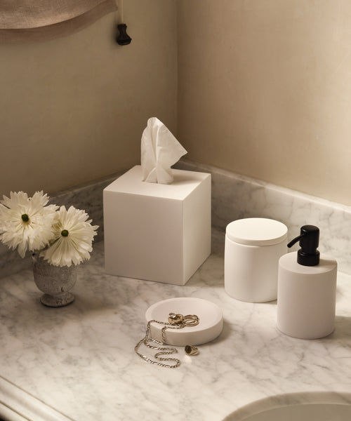 A marble bathroom countertop displays elegant home accessories: white containers, the Coast Tissue Cover, a soap dispenser, a small vase with white flowers, and jewelry including a chain necklace and rings.