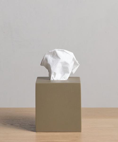 A beige Coast Tissue Cover, crafted from handcasted resin, rests on a light wooden surface with a crumpled white tissue partially pulled from the top against a plain light gray background.
