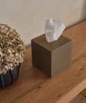 A square Coast Tissue Cover in olive green sits on a wooden surface beside dried hydrangeas, with a white tissue pulled up. This stylish accessory adds charm to your home décor against a beige background wall.