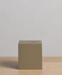 The Coast Tissue Cover, a simple, matte olive-green cube crafted from handcasted resin, sits on a light wooden surface against a plain light gray background.