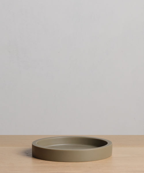 The Coast Tray, a round and shallow olive-green dish crafted from handcast resin, sits centered on a light wooden surface against a plain gray background—adding modern style to your home accessories collection.