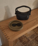 A small black handbag and black sunglasses sit on the Coast Tray, a round olive-green handcast resin accessory, placed atop a wooden table with dovetail joints, next to a tiled floor.
