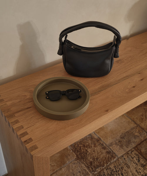 A small black handbag and black sunglasses sit on the Coast Tray, a round olive-green handcast resin accessory, placed atop a wooden table with dovetail joints, next to a tiled floor.