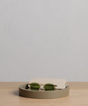 A pair of green-lensed sunglasses with a beige case rests in a round, shallow Coast Tray on a light wooden surface against a plain, light-colored background.