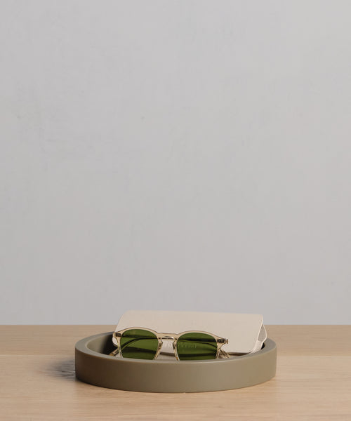A pair of green-lensed sunglasses with a beige case rests in a round, shallow Coast Tray on a light wooden surface against a plain, light-colored background.