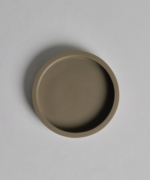 The handcasted resin Coast Tray, round and flat with a taupe finish, sits empty at the center of a light gray textured surface, elegantly displayed from above.