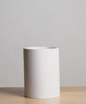 A plain white ceramic cylinder vase sits on a light wooden surface against a neutral gray background, ideal for pairing with accessories such as the Coast Waste Basket.