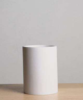 A plain white ceramic cylinder vase sits on a light wooden surface against a neutral gray background, ideal for pairing with accessories such as the Coast Waste Basket.