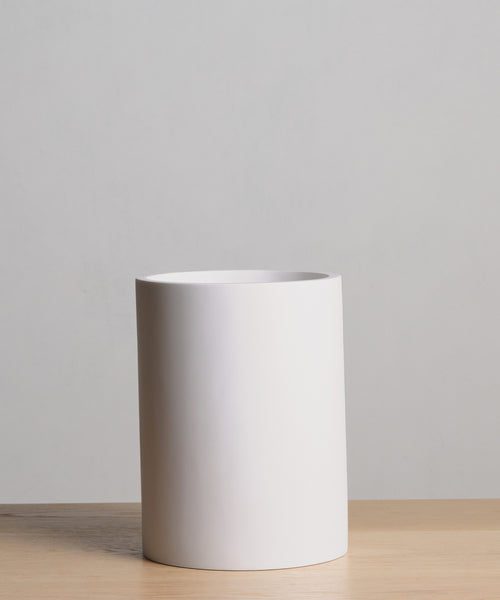 A plain white ceramic cylinder vase sits on a light wooden surface against a neutral gray background, ideal for pairing with accessories such as the Coast Waste Basket.