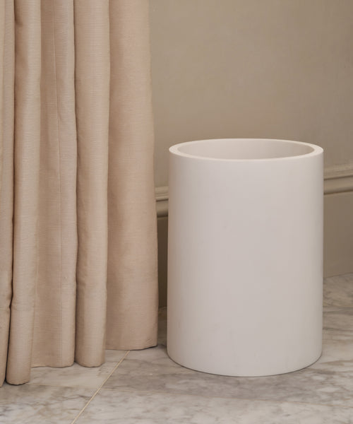 The Coast Waste Basket, crafted from handcasted resin in plain white, adds an elegant touch next to beige curtains and a pale wall on a marble floor—perfect for elevating your home accessories.