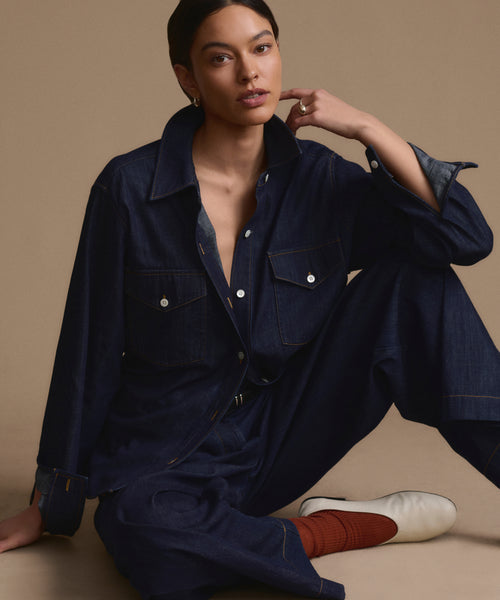 A woman with short dark hair sits on the floor against a beige background, wearing the Western-inspired Colt Shirt and matching denim pants, white shoes, and rust socks. She looks at the camera, one hand resting near her chin.