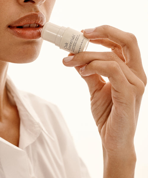 A person in a white shirt gently applies Conditioning Lip Balm to their lips, holding it with one hand against a neutral background.