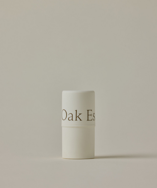A white cylindrical tube of Conditioning Lip Balm with a minimal design stands upright against a plain beige background. The container features the text Oak Es.