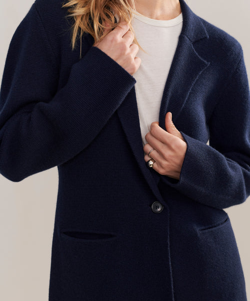 A person wearing the Cooper Blazer in dark navy over a white top, holding the lapel with one hand and adjusting a button with the other. Their face is not visible.