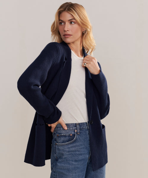 A woman with wavy blonde hair stands against a plain background, looking to the side. She wears the Cooper Blazer in dark navy wool cashmere over a white tee and blue jeans, with one hand in her pocket.