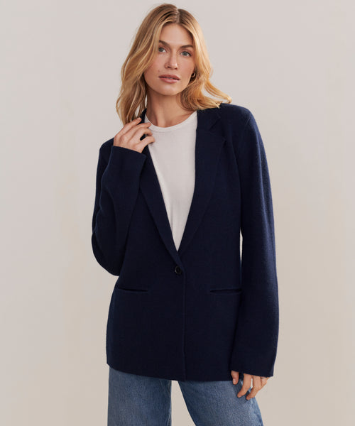 A woman with blonde hair models the Cooper Blazer in dark navy over a white top and blue jeans, standing against a neutral background and looking at the camera with a relaxed expression.