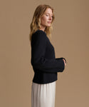 A woman in side profile wearing a black knit sweater and a light cream skirt, set against a solid warm beige studio background.