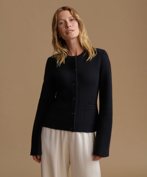A woman stands against a plain beige studio background wearing a black cardigan with front buttons, off-white wide-leg trousers.