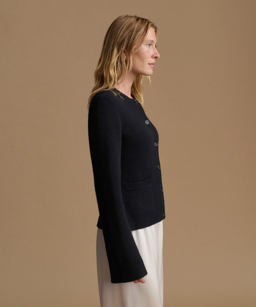 A woman shown in side profile wearing a black long-sleeve button-front cardigan with patch pockets and white wide-leg pants, set against a solid warm beige studio background.
