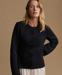 A woman wearing a black knit cardigan sweater with front buttons and patch pockets, paired with a cream-colored skirt, set against a solid warm brown studio background.
