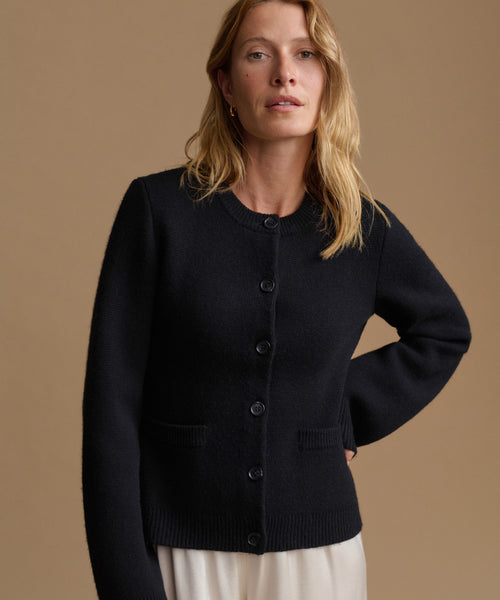 A woman wearing a black knit cardigan sweater with front buttons and patch pockets, paired with a cream-colored skirt, set against a solid warm brown studio background.