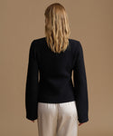 A woman viewed from behind wearing a black long-sleeve knit sweater and cream-colored trousers, set against a solid warm beige studio background.