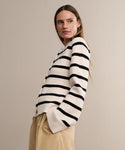 A woman with long hair stands sideways against a plain beige background, looking over her shoulder while wearing the Cooper Cardigan in ivory black stripes paired with beige pants.