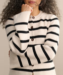 A person with curly hair wears the Cooper Cardigan in ivory black stripe with brown buttons, crossing their arms and thoughtfully touching their chin.