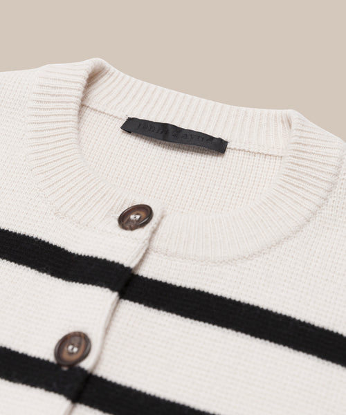 The Cooper Cardigan is a cream knit with ivory and black stripes, ribbed collar, brown buttons, and a black label inside the neckline. Its structured silhouette contrasts against the light beige background.