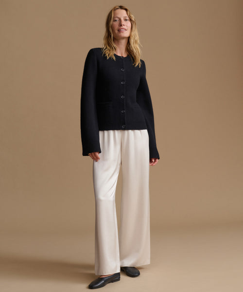 A woman stands against a plain beige studio background wearing a black cardigan with front buttons, off-white wide-leg trousers, and dark flat shoes.