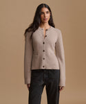 A woman modeling a light beige knit cardigan with dark buttons, paired with dark jeans, against a solid beige studio background.