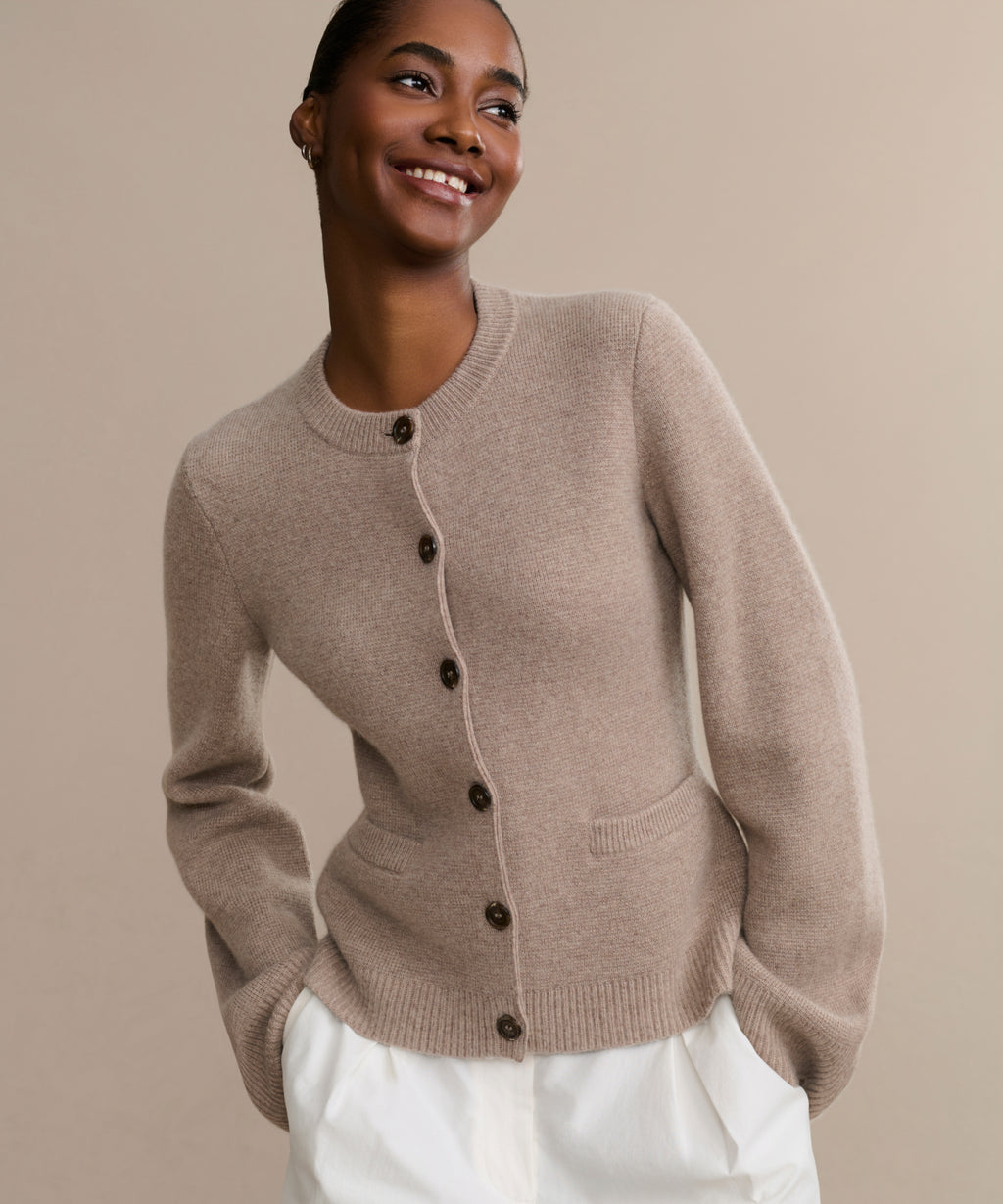 A smiling person stands before a beige backdrop, modeling the Cooper Cardigan with front pockets and white pants, showcasing its sleek silhouette for versatile looks.
