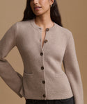 A woman wearing a light taupe knit cardigan sweater with dark brown buttons, shown from the chest up against a solid warm beige background.