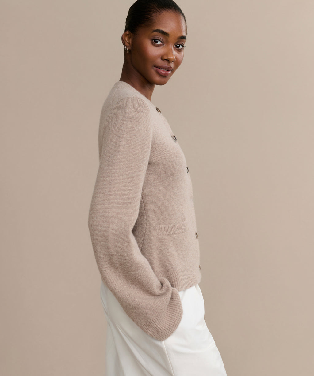 A woman stands sideways, facing the camera, wearing the Cooper Cardigan—featuring a structured silhouette and front pockets—paired with white pants against a plain beige background.
