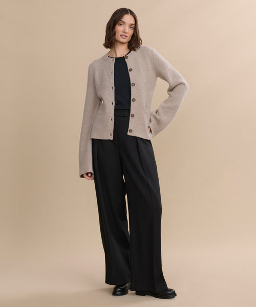 A woman with shoulder-length brown hair poses confidently against a beige background, wearing the Cooper Cardigan in taupe, black wide-leg pants, and black shoes, with one hand on her hip.