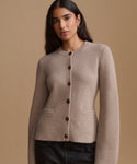 A woman wearing a light taupe knit cardigan sweater with dark buttons, paired with dark jeans, set against a solid warm beige background.