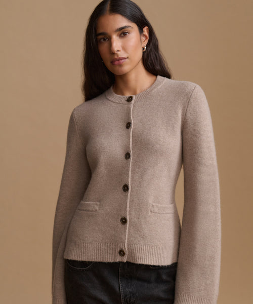 A woman wearing a light taupe knit cardigan sweater with dark buttons, paired with dark jeans, set against a solid warm beige background.