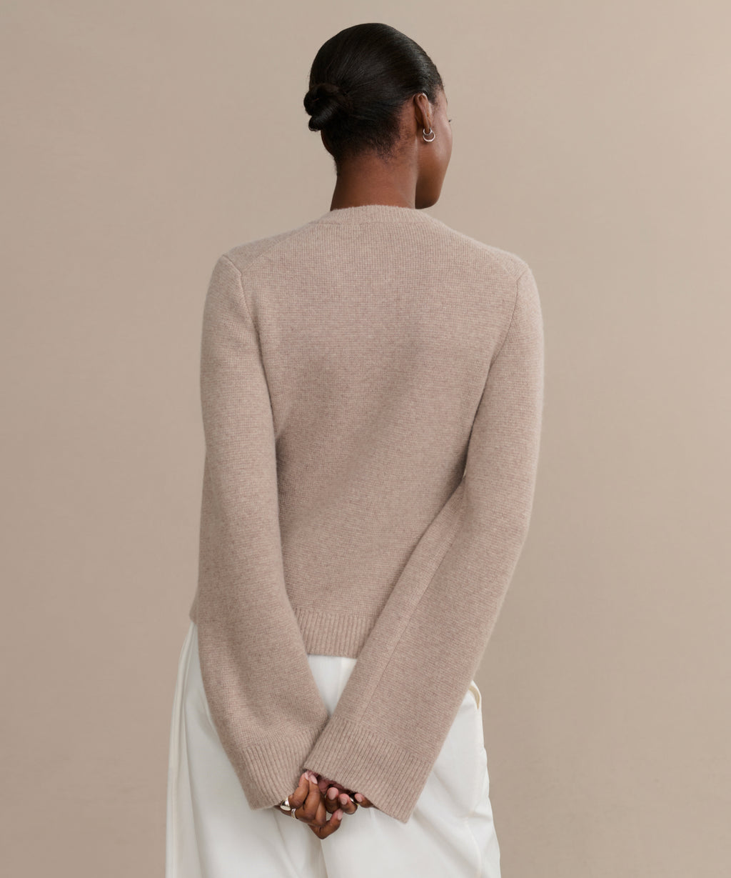 Wearing the Cooper Cardigan and a white skirt, a person with short, neat hair stands with hands behind their back against a plain beige background, emphasizing the cardigan’s structured silhouette and versatile style.