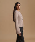 A woman wearing a light beige knit cardigan with dark buttons, shown in profile against a solid tan studio background.