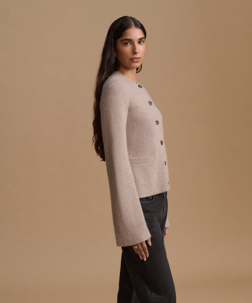 A woman wearing a light beige knit cardigan with dark buttons, shown in profile against a solid tan studio background.
