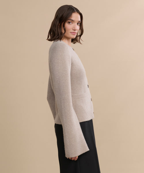 A woman with shoulder-length brown hair wears the Cooper Cardigan, known for its structured silhouette and wide sleeves, paired with a black skirt, standing in profile against a plain beige background.