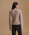 A woman shown from the back wearing a light taupe knit sweater and dark jeans, standing against a solid warm beige studio background.