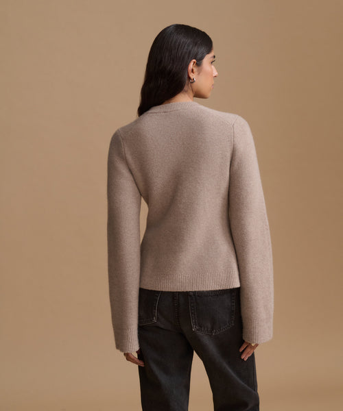 A woman shown from the back wearing a light taupe knit sweater and dark jeans, standing against a solid warm beige studio background.